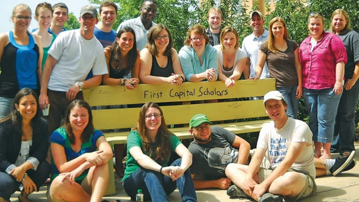 Students gather around a bench that says, "First Capital Scholars."