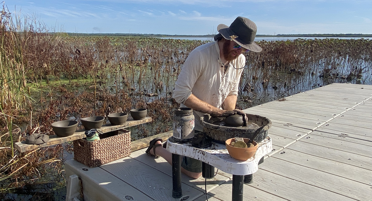 A person shapes clay on a pottery wheel outdoors on a dock at the Emiquon Preserve, surrounded by wetland plants and water.