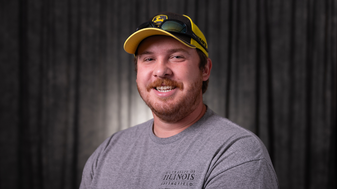 Nathan Curry, a smiling man in a yellow cap and gray shirt against a dark background.