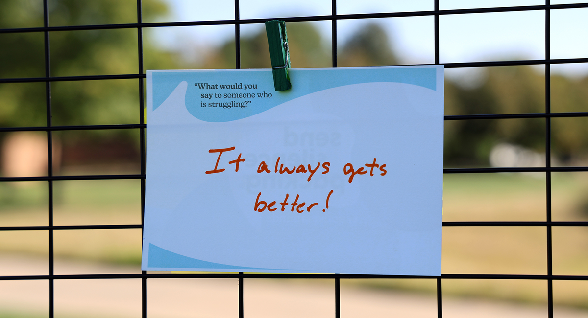 Note with "It always gets better!" clipped to a fence, blurry campus in background.