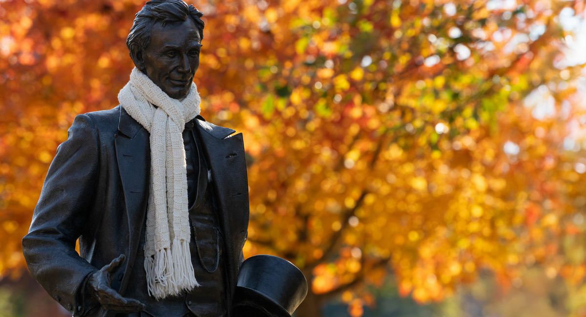 the lincoln statue on the quad has a real scarf wrapped around his neck and there are autumnal leaves on a tree in the background