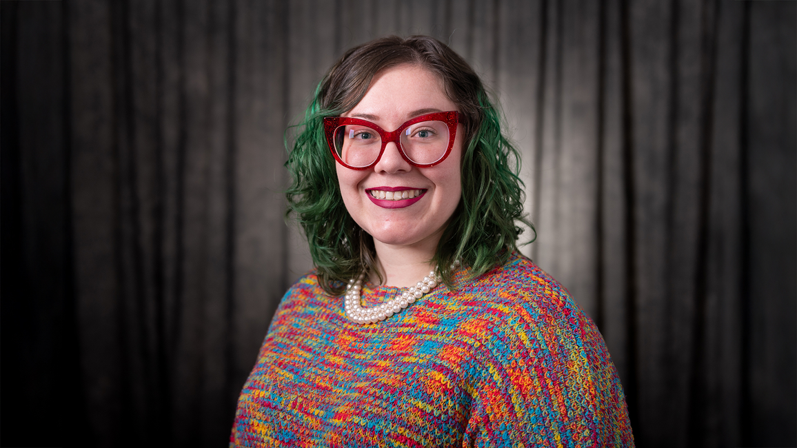 Dre Duvendack Visk with glasses, colorful sweater, dark curtain background, smiling.
