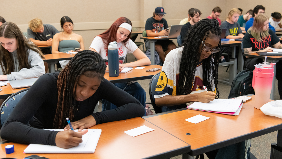 Students attentively writing in a classroom, sitting at desks with notebooks.