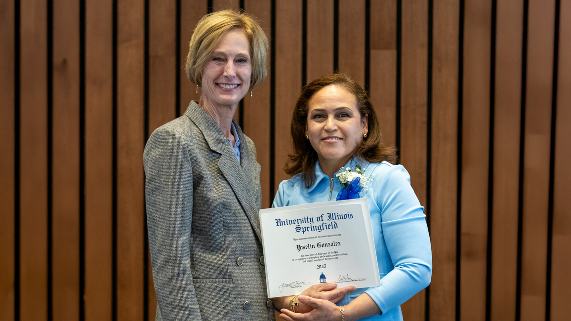 Two women smiling, one holds a certificate, standing against a wood-paneled wall.
