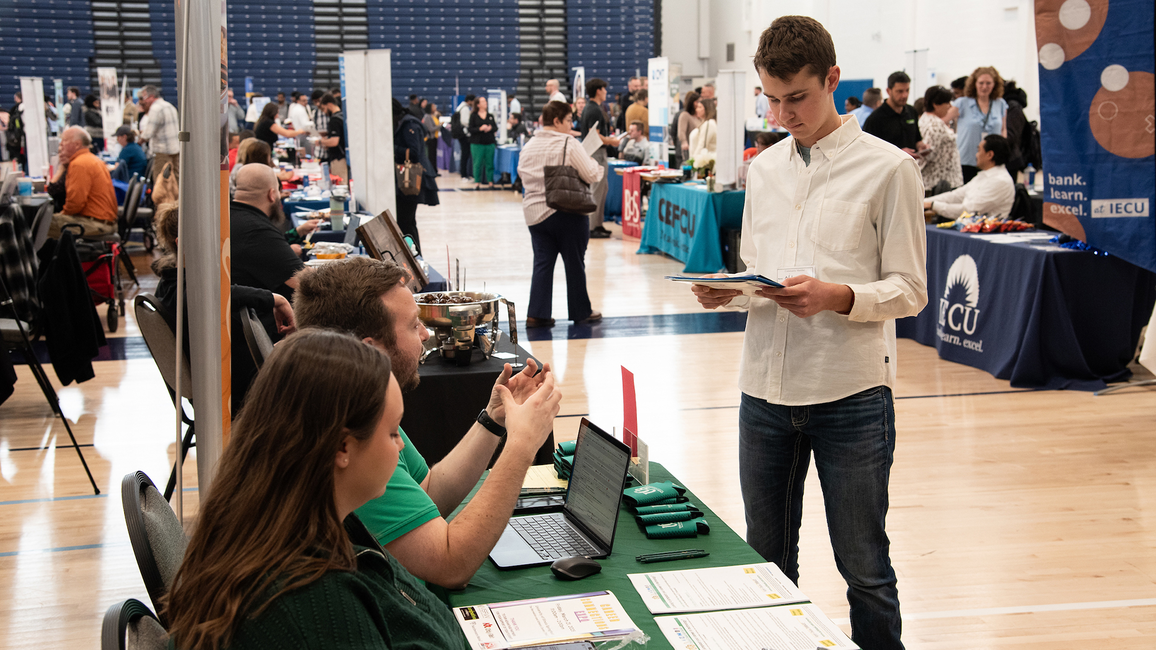 Man in white shirt talks to seated people at a busy job fair.