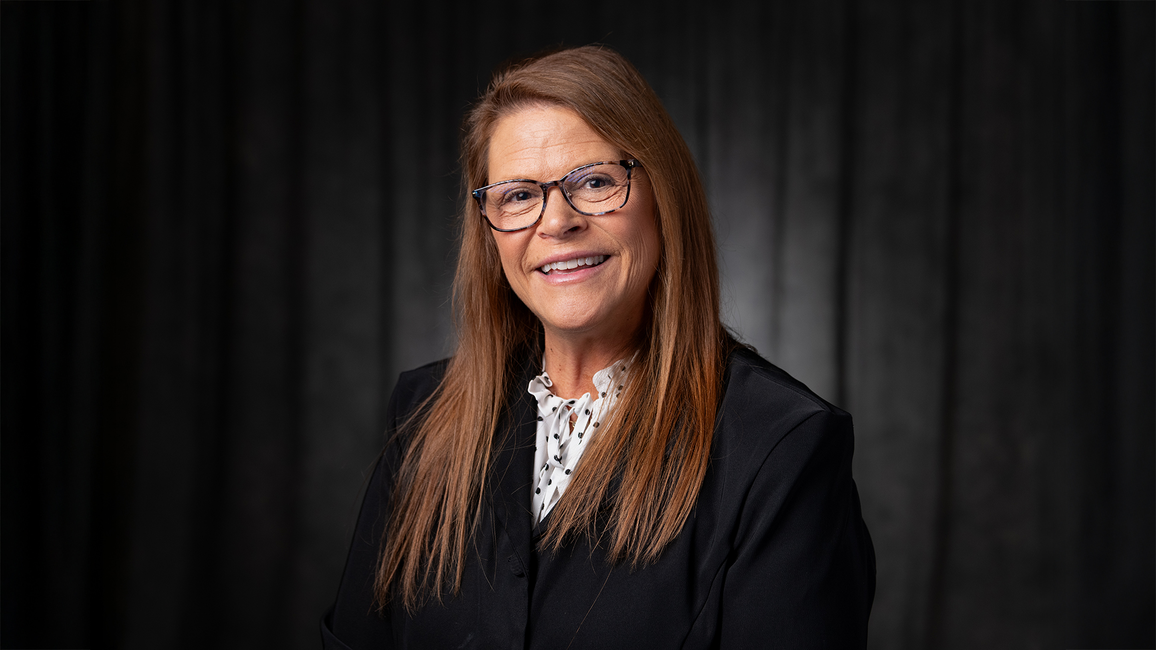 Smiling woman in a dark suit and patterned blouse, against a black background.