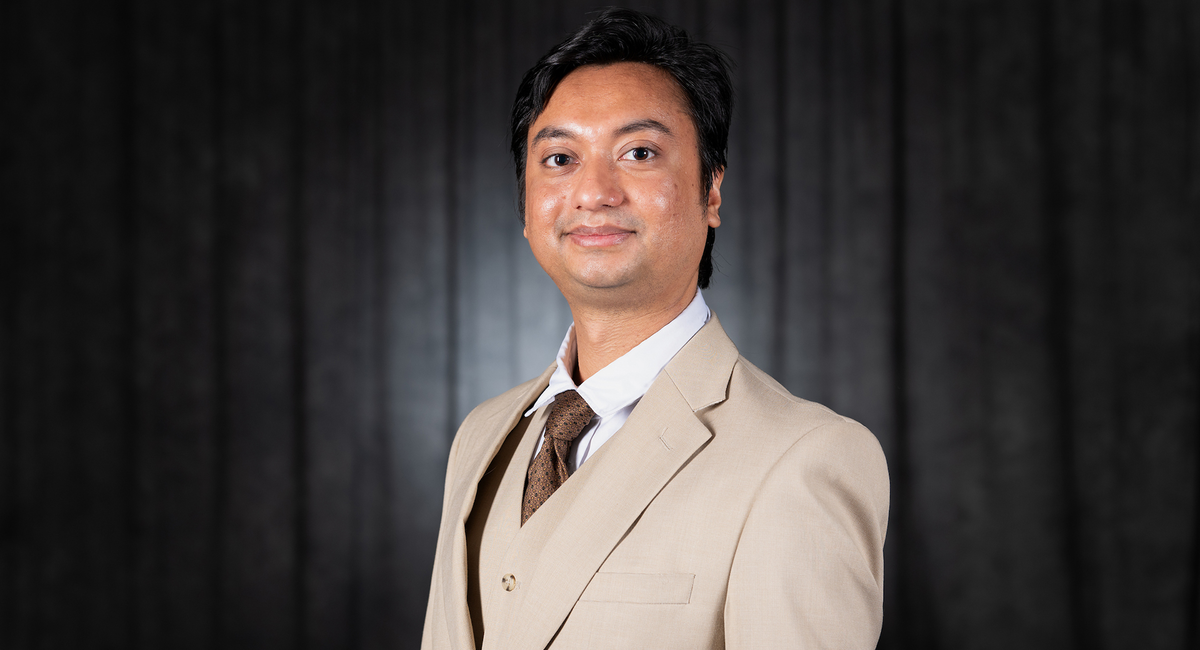 A man in a beige suit and tie, smiling, against a dark background.