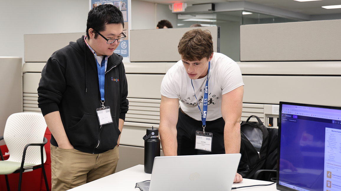 Two men discussing work at a desk with a laptop in an office setting.