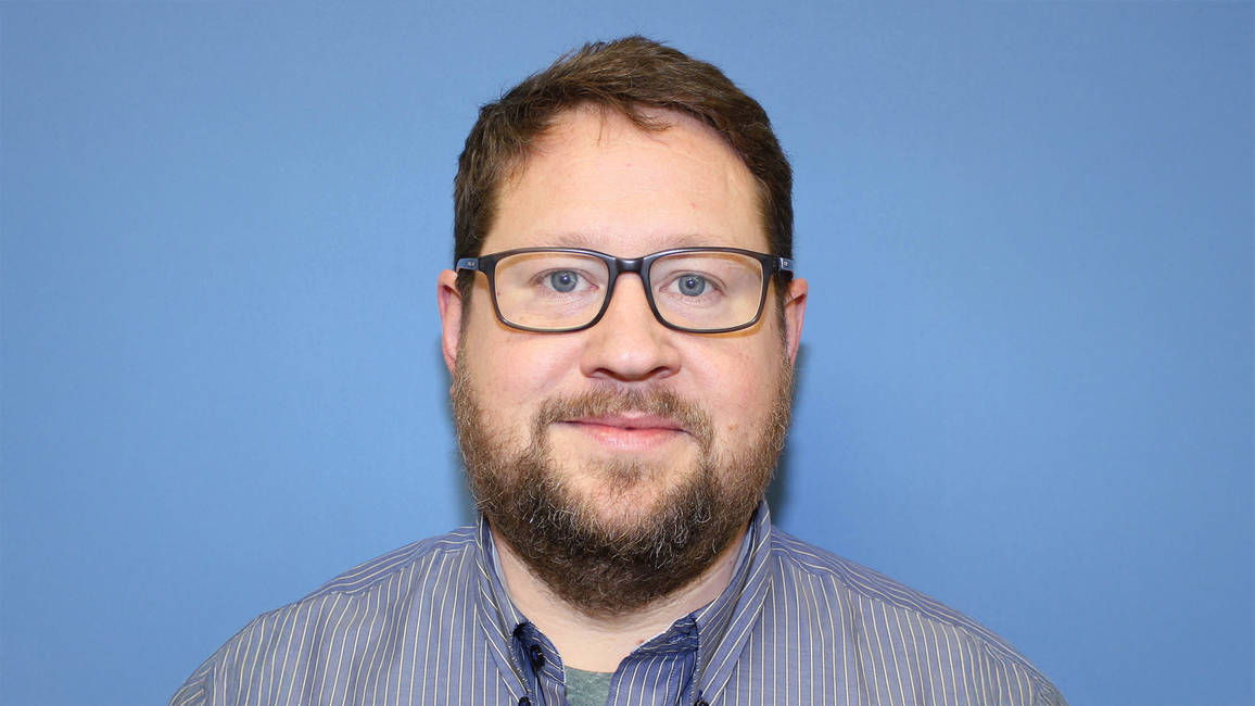 Jeff Sudduth, a man with short brown hair, beard and glasses wearing a striped shirt, photographed against a plain blue background.