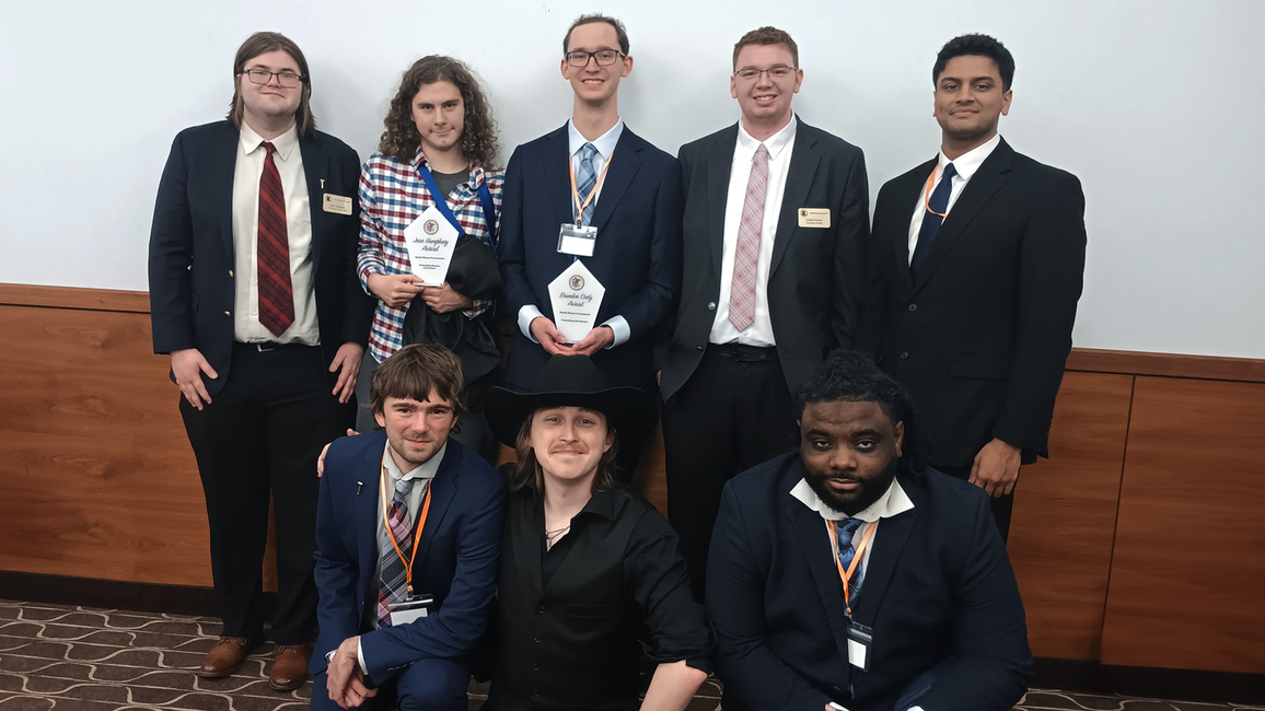 Group of people in formal attire, two holding awards, smiling for a photo indoors.