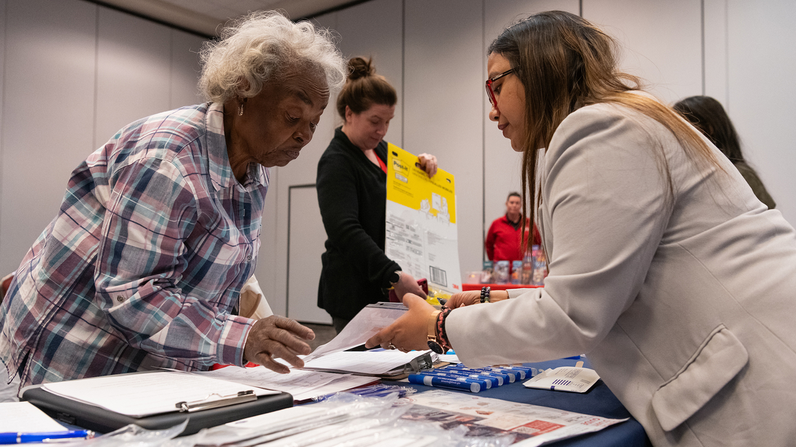 Two women exchanging documents at a crowded conference table.