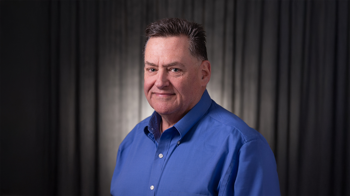 Man in a blue shirt against a dark curtain background, smiling slightly.
