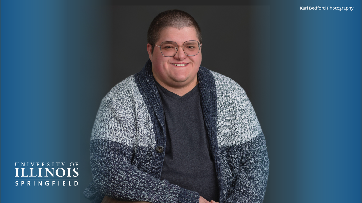 Smiling person in glasses and a blue-gray sweater against a dark background.