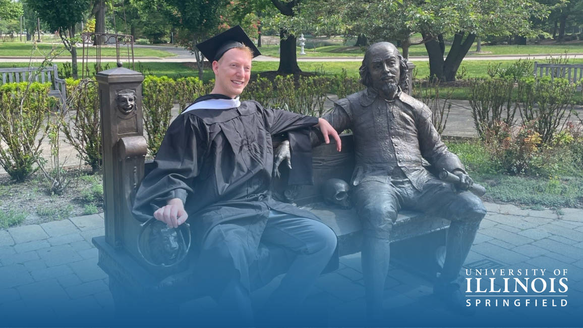 Graduate sitting next to a statue on a bench in a park setting, smiling.