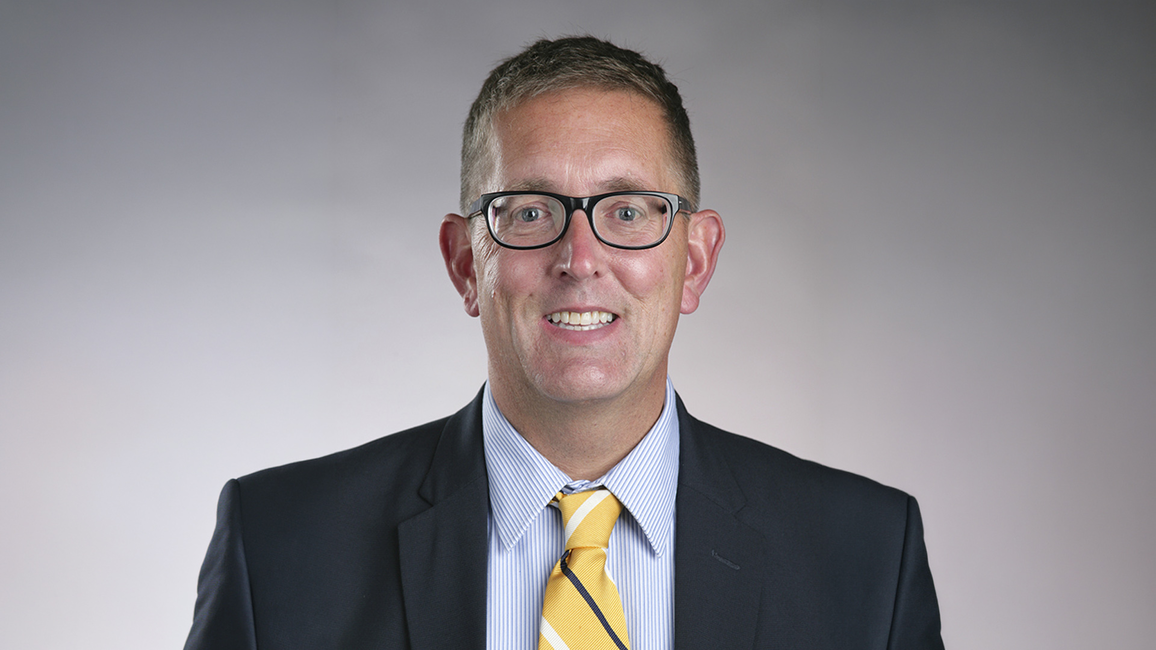 Man in glasses and suit with yellow tie, smiling against a dark gradient background.