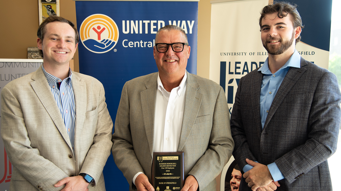 Three men in suits stand smiling, one holding a plaque, with banners in the background.