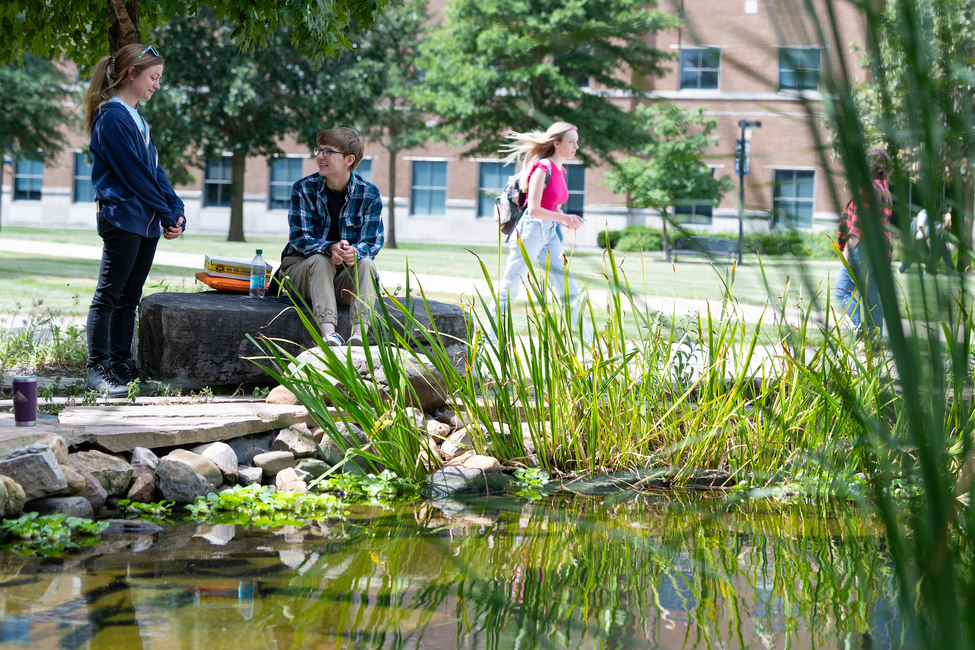 Students by a pond on a sunny day, university building in background.