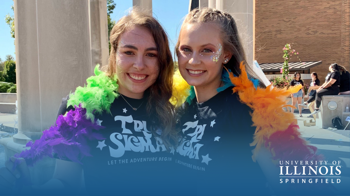 Two people smiling, wearing rainbow feather boas outdoors in sunlight. Their t-shirts say Tri Sigma.