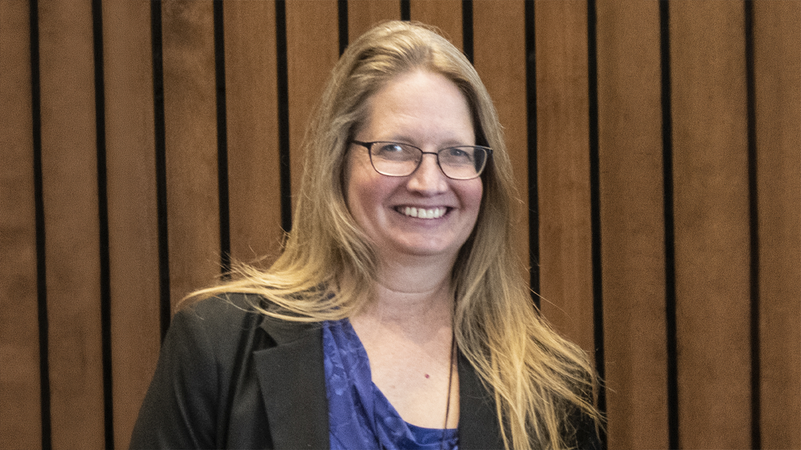 Smiling woman with long hair, wearing glasses, black blazer, blue top. Brown striped background.