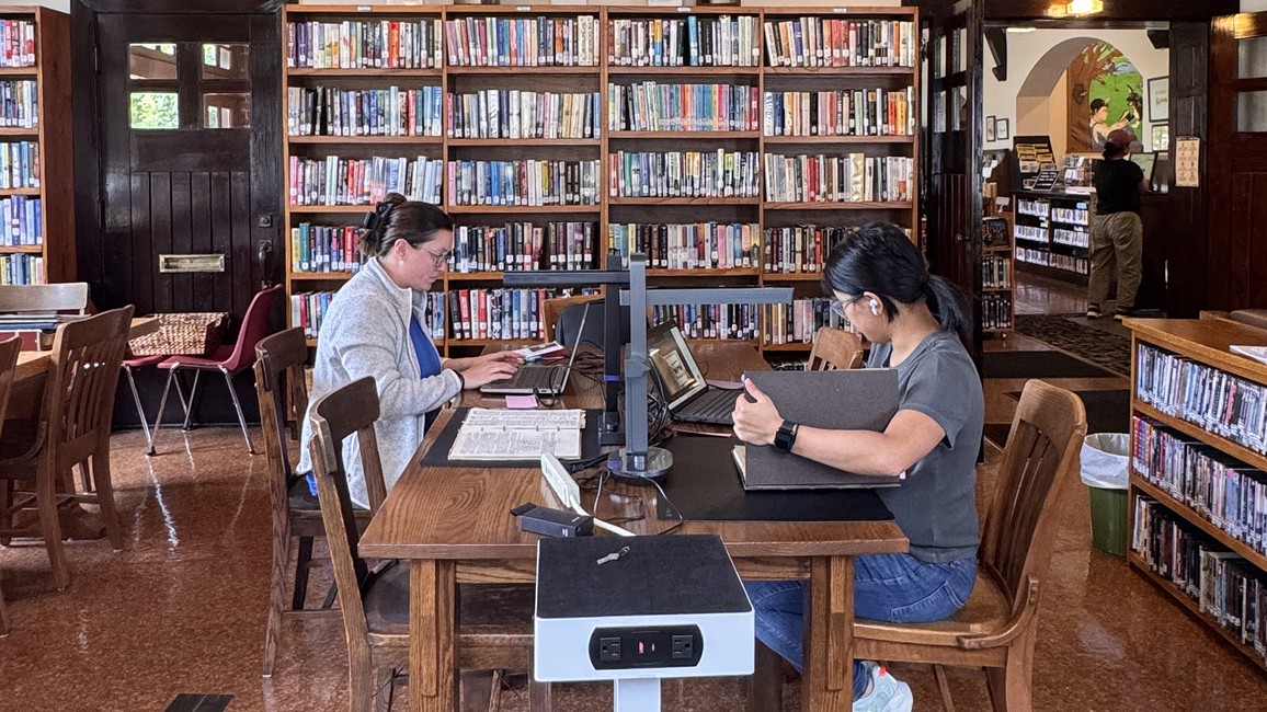 Library with two people working at a wooden table, surrounded by bookshelves.