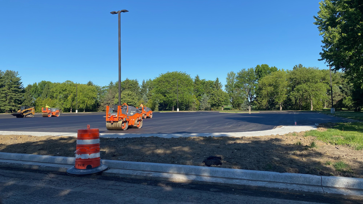 Freshly paved parking lot with orange traffic barrels and trees in the background.