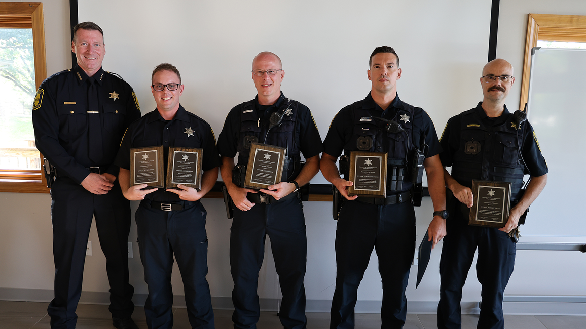 Five men in uniform holding plaques, standing indoors, smiling.