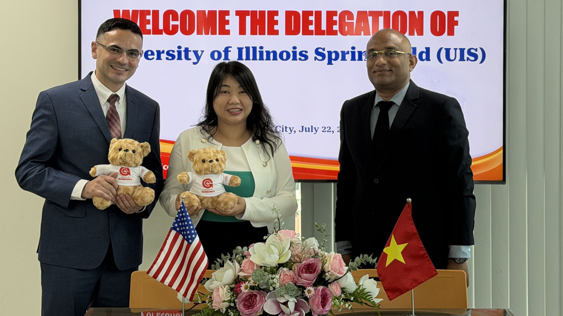 Three people smiling, holding teddy bears, with flags and flowers on a table.