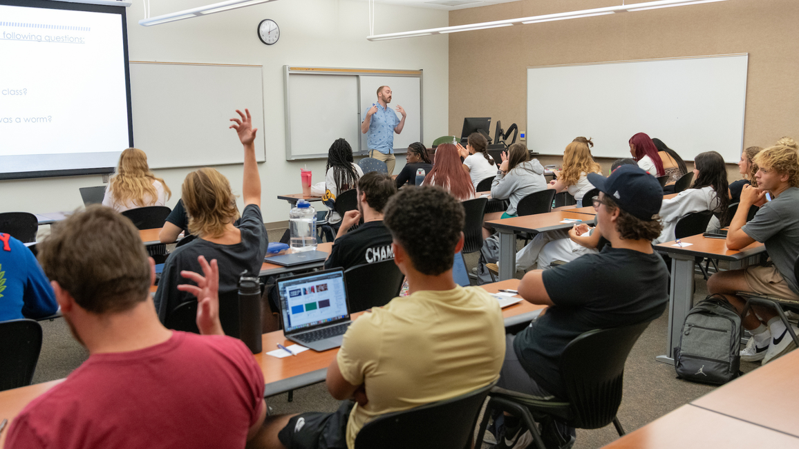 Classroom with students, one raising hand, and teacher at the front near a projector screen.