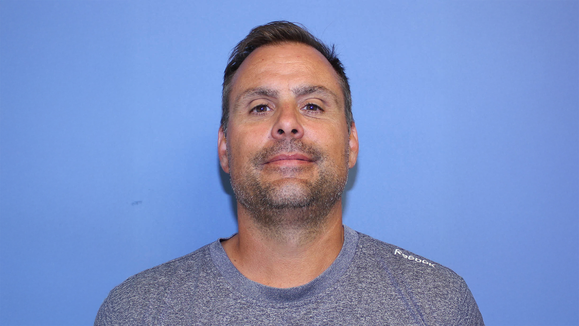 Man in a gray shirt against a blue background, smiling slightly.