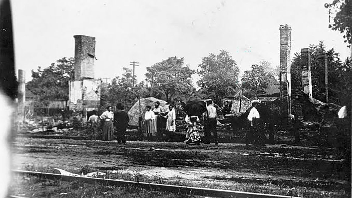 Group standing near train tracks, trees, and ruins in black and white photograph.