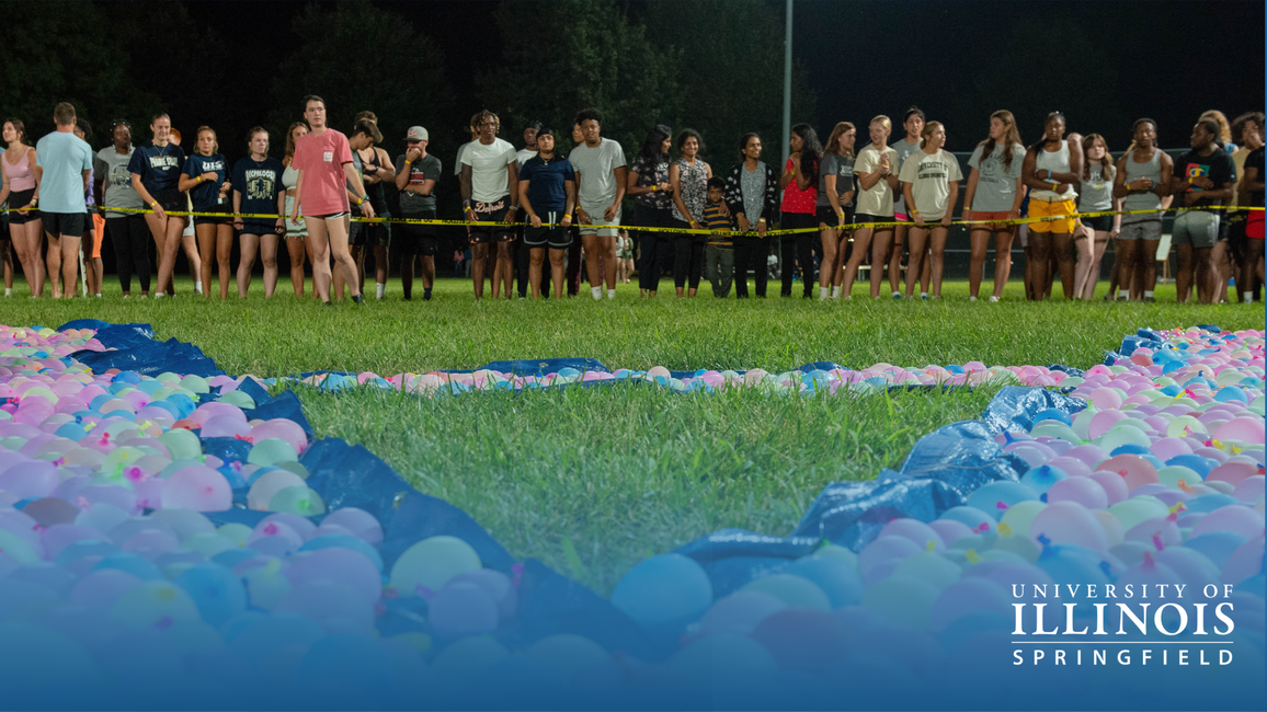 Crowd gathers at night on grass, with colorful water balloons in foreground.