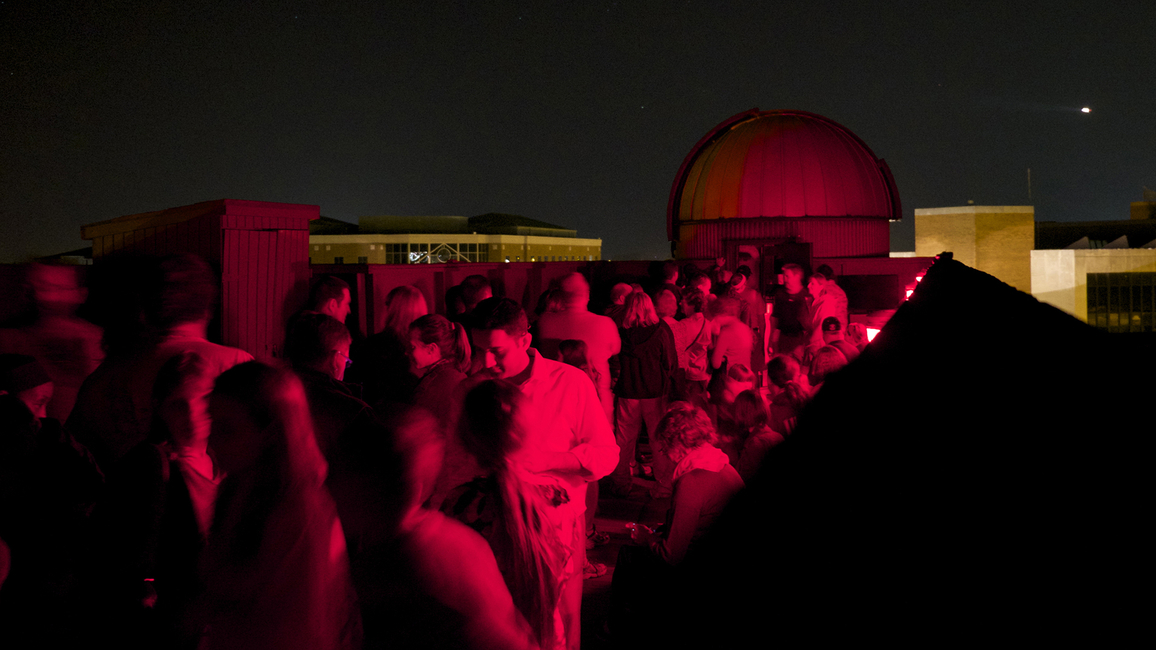 Crowd gathered on a rooftop at night, illuminated by red lights.
