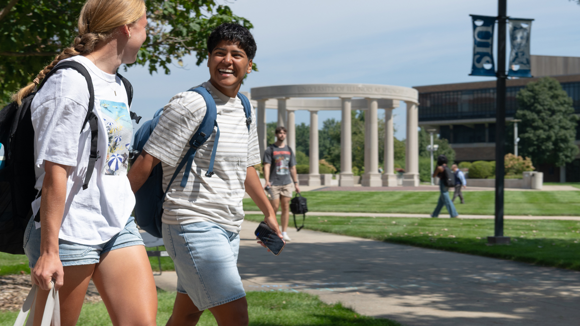 Two students walking on a campus path, smiling, with a stone pavilion in the background.