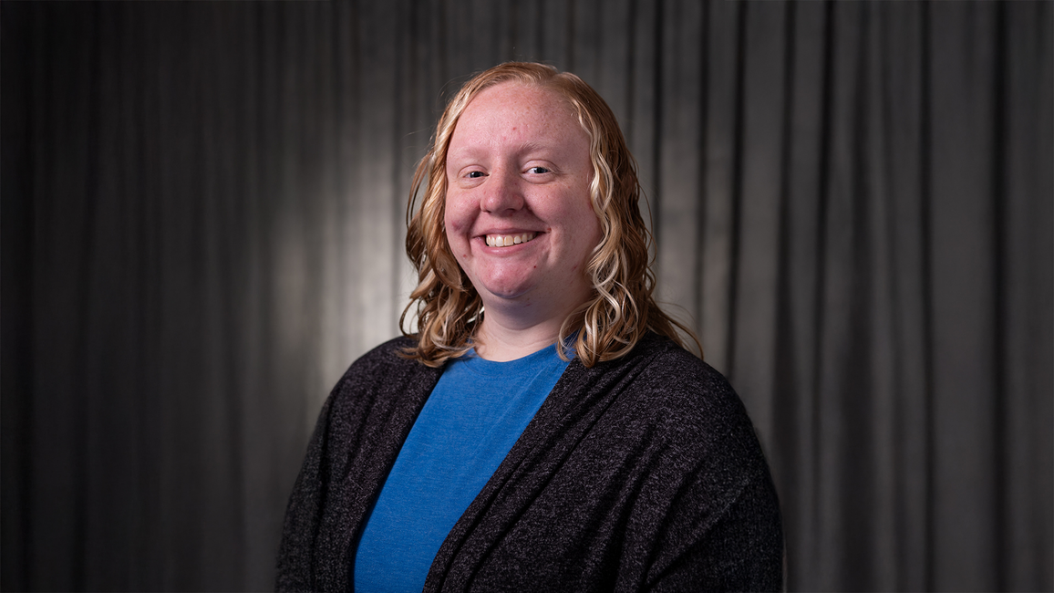 Smiling person with long hair against a dark curtain background.