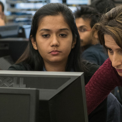 a professor standing next to a student at a computer desk showing the student something on a computer