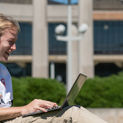 a young man sitting on the quad laughing while looking at his laptop
