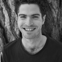 Smiling man in black-and-white, standing against tree bark.