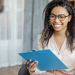 Woman with glasses smiling, holding clipboard, sitting across from another person.