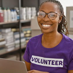 Smiling volunteer in a purple shirt, working on a laptop in a storage room.