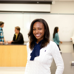 a female student smiling in a classroom setting with a professor and students in the background