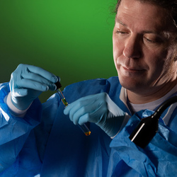 Scientist in blue protective gear under green lighting using a dropped to add liquid in beaker.