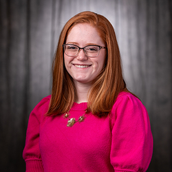Smiling person with red hair and glasses, wearing a pink sweater, against a dark backdrop.