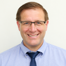 Smiling man wearing glasses, blue shirt, and navy tie.
