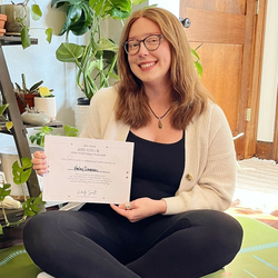 Haley Simpson holding a yoga certification while sitting on a yoga mat with plants in the background.