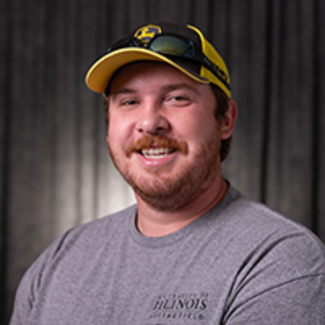 Nathan Curry, a smiling man in a yellow cap and gray shirt against a dark background.