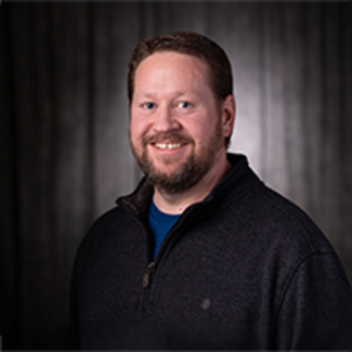 Jeff Sudduth, a smiling man in dark sweater against a dark background.