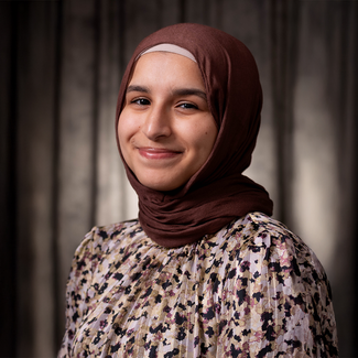 Smiling person in a floral blouse and dark hijab in front of a grey background.