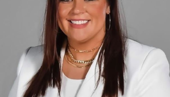 Summer Quesenberry portrait in a white blazer and white top, gold necklaces, long dark hair.