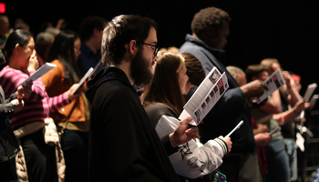 People standing and singing from sheets in a concert hall.
