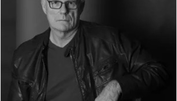 Man in a leather jacket, sitting in dim lighting, looking serious.